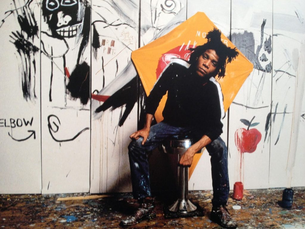 Jean-Michel Basquiat sitting in front of a wall with his paining